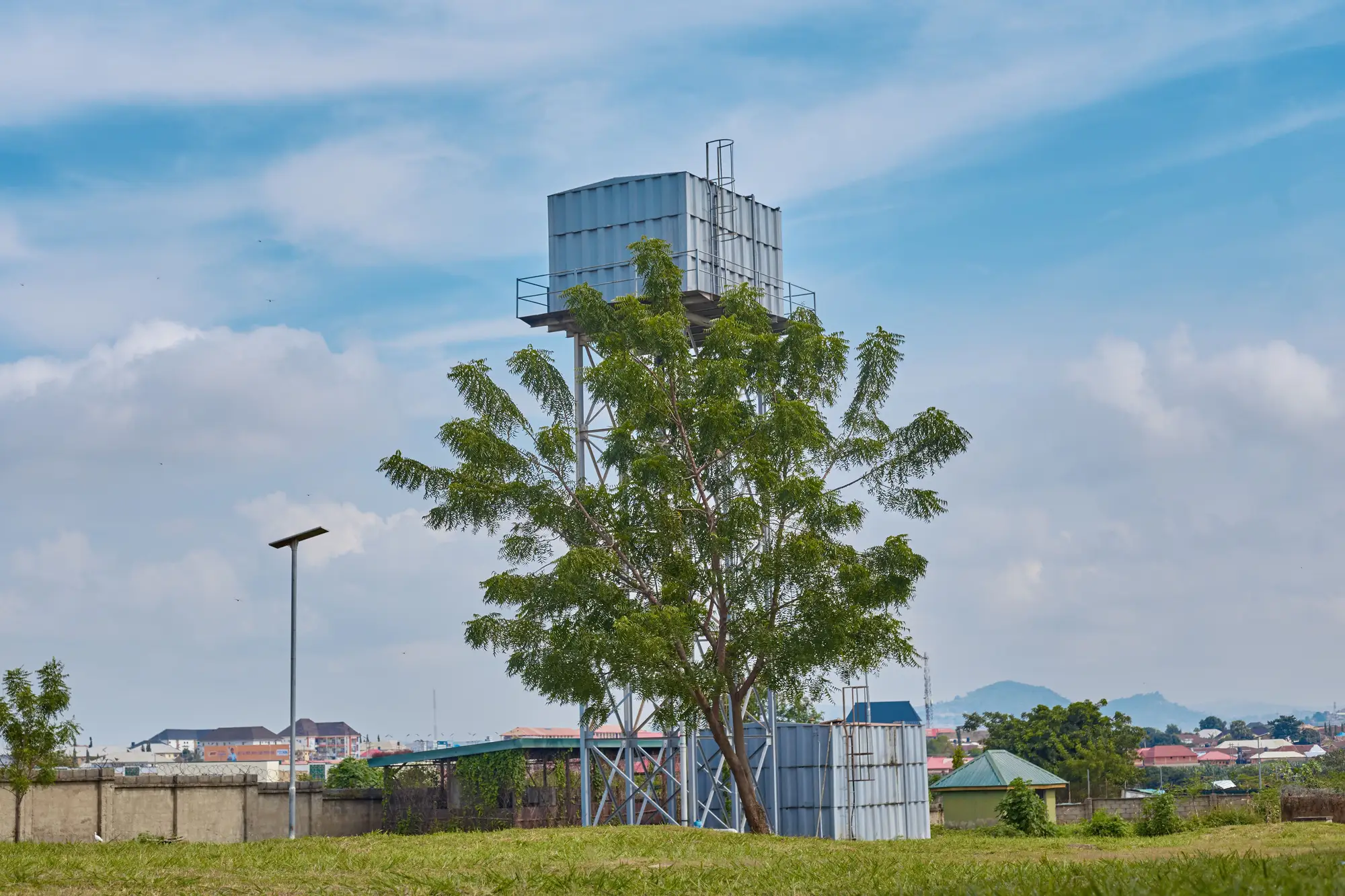 A storage tank on a tall steel stand behind a tree, at the edge of a town